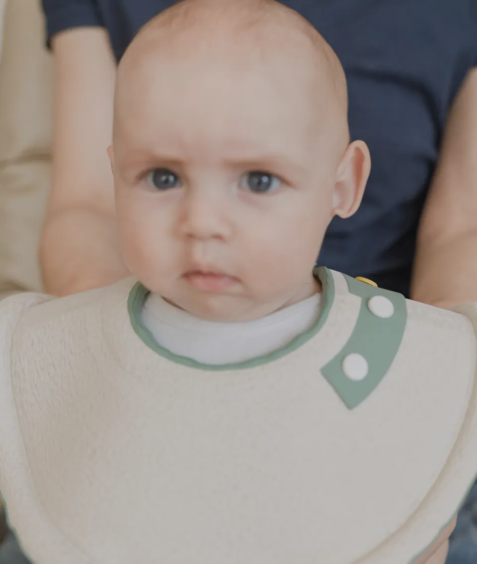 Baby wearing the mybetterbib design, with a wide ivory absorbent shelf and sage-trimmed closure